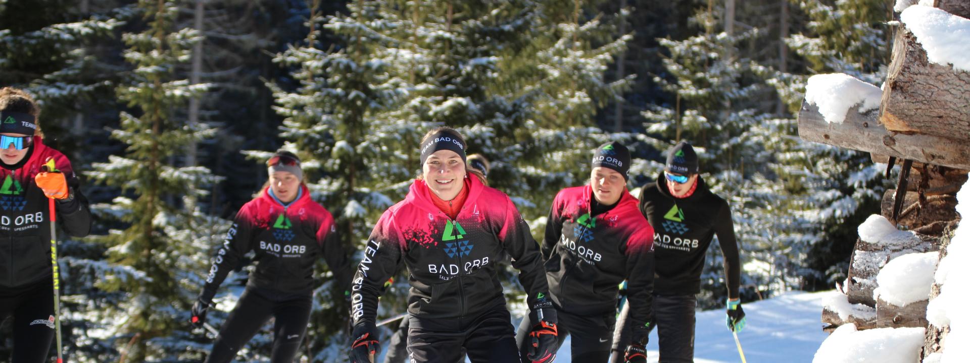 Mehrere Skilangläuferinnen und Skilangläufer fahren im seleben Team-Outfit über eine verschneite Langlauf-Loipe