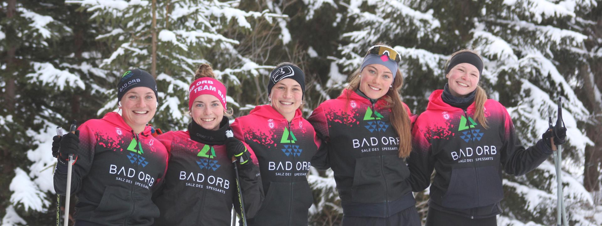 Fünf junge Sportlerinnen im gleichen Team-Dress posieren lächelnd mit Langlaufstöcken in der Hand für ein Gruppenbild vor verschneiten Tannen