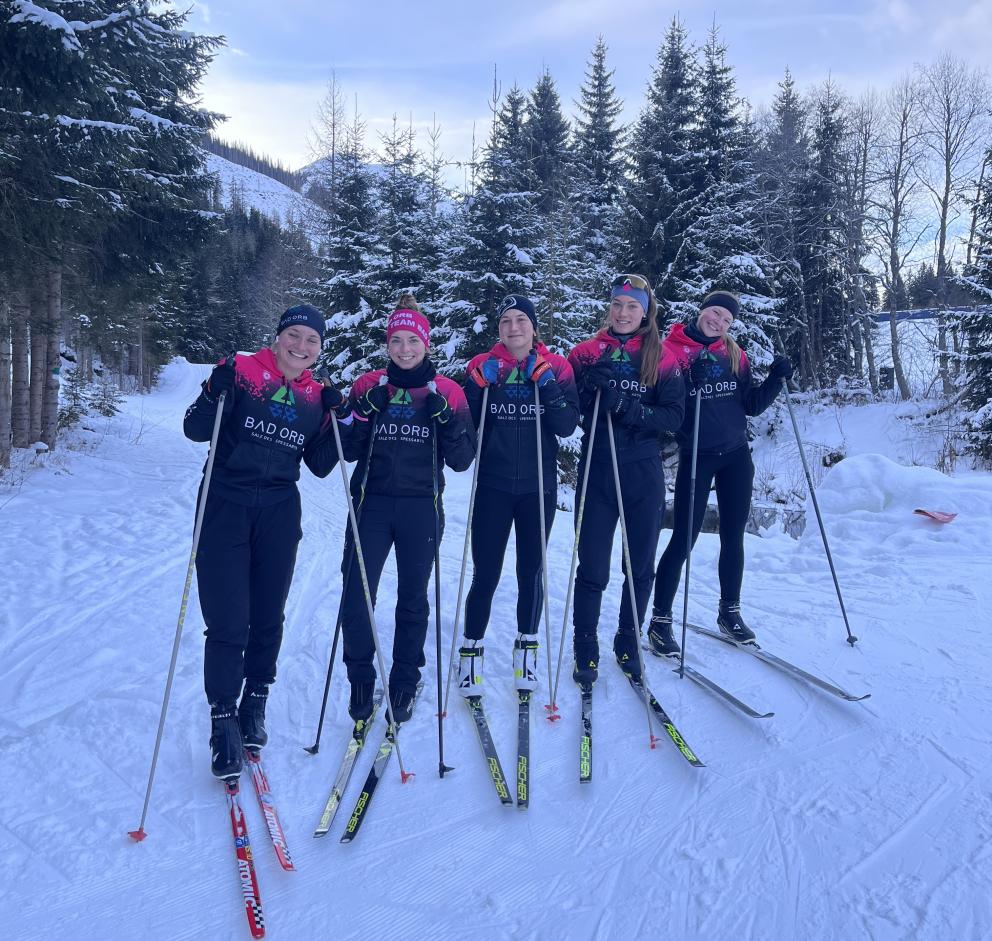 Fünf junge Sportlerinnen posieren - im gleiche Team-Outfit gekleidet - auf Langlaufskiern in einer Winterlandschaft für ein Gruppenbild