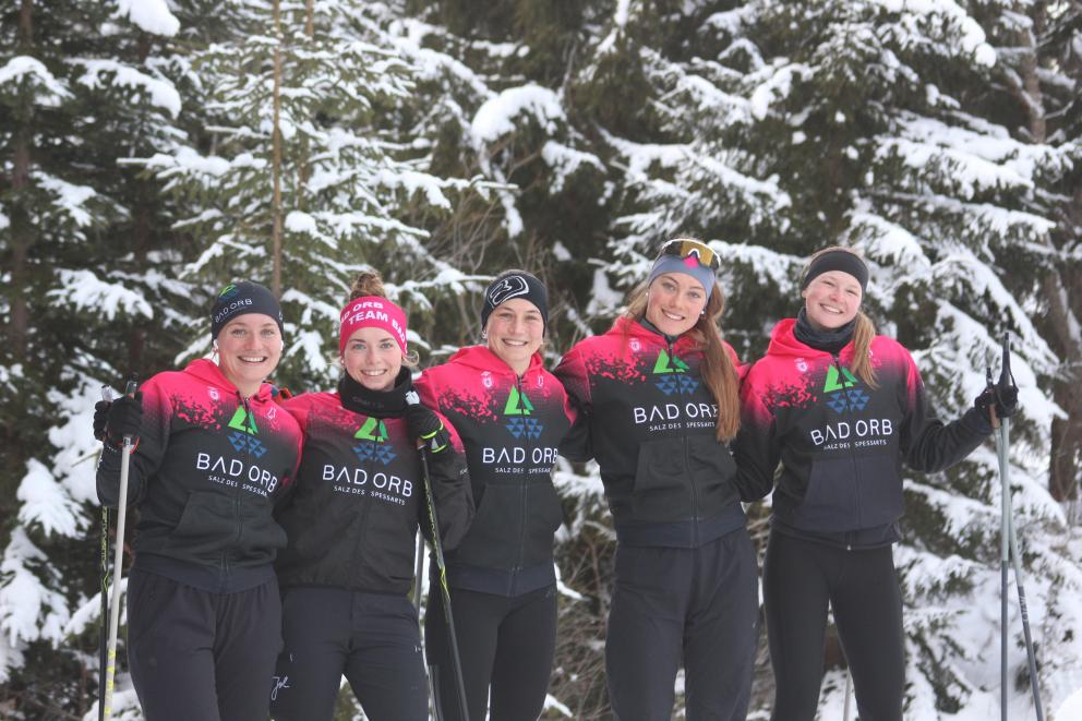 Fünf junge Sportlerinnen im gleichen Team-Dress posieren lächelnd mit Langlaufstöcken in der Hand für ein Gruppenbild vor verschneiten Tannen