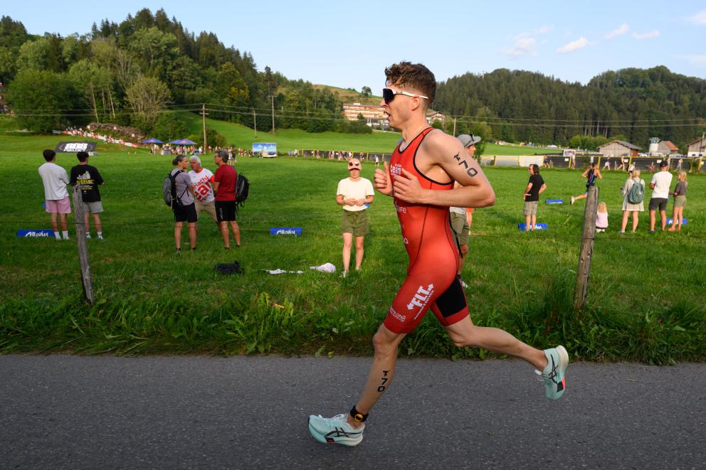 Mann in orangenen Einteiler läuft an einer Wiese vorbei, auf der Zuschauer sind