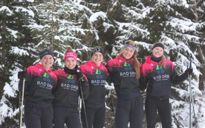 Fünf junge Sportlerinnen im gleichen Team-Dress posieren lächelnd mit Langlaufstöcken in der Hand für ein Gruppenbild vor verschneiten Tannen
