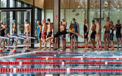 Ein Schwimmer im kurzen Neoprenanzug springt vom Sprungblock ins Wasser. Dahinter stehen weitere männliche Sportler, die ebenfalls in der Halle in einem Wettkampf antreten und in Reihe auf ihren Start warten